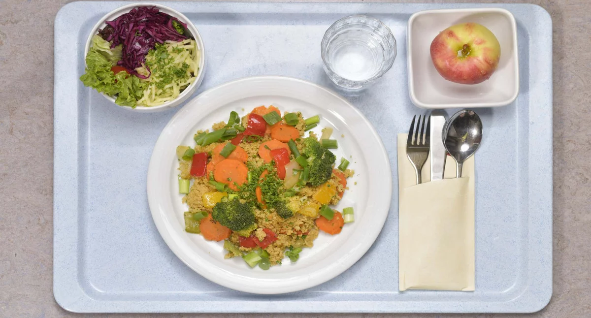 Auf einem Tablett ist ein buntes Mittagessen mit Salat, Apfel, Wasserglas und Besteck abgebildet.