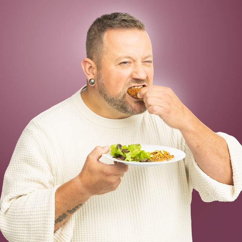 Ein Mann hält in der rechten Hand einen Teller mit Blattsalat und beißt herzhaft in eine Frikadelle.