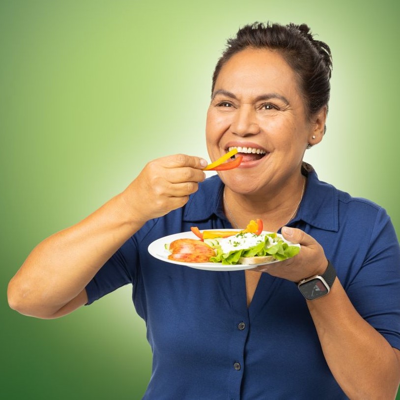 Eine Frau hält lächelnd einen Teller mit Gemüsestreifen auf der linken Hand und führt zwei Paprikastreifen zum Mund.